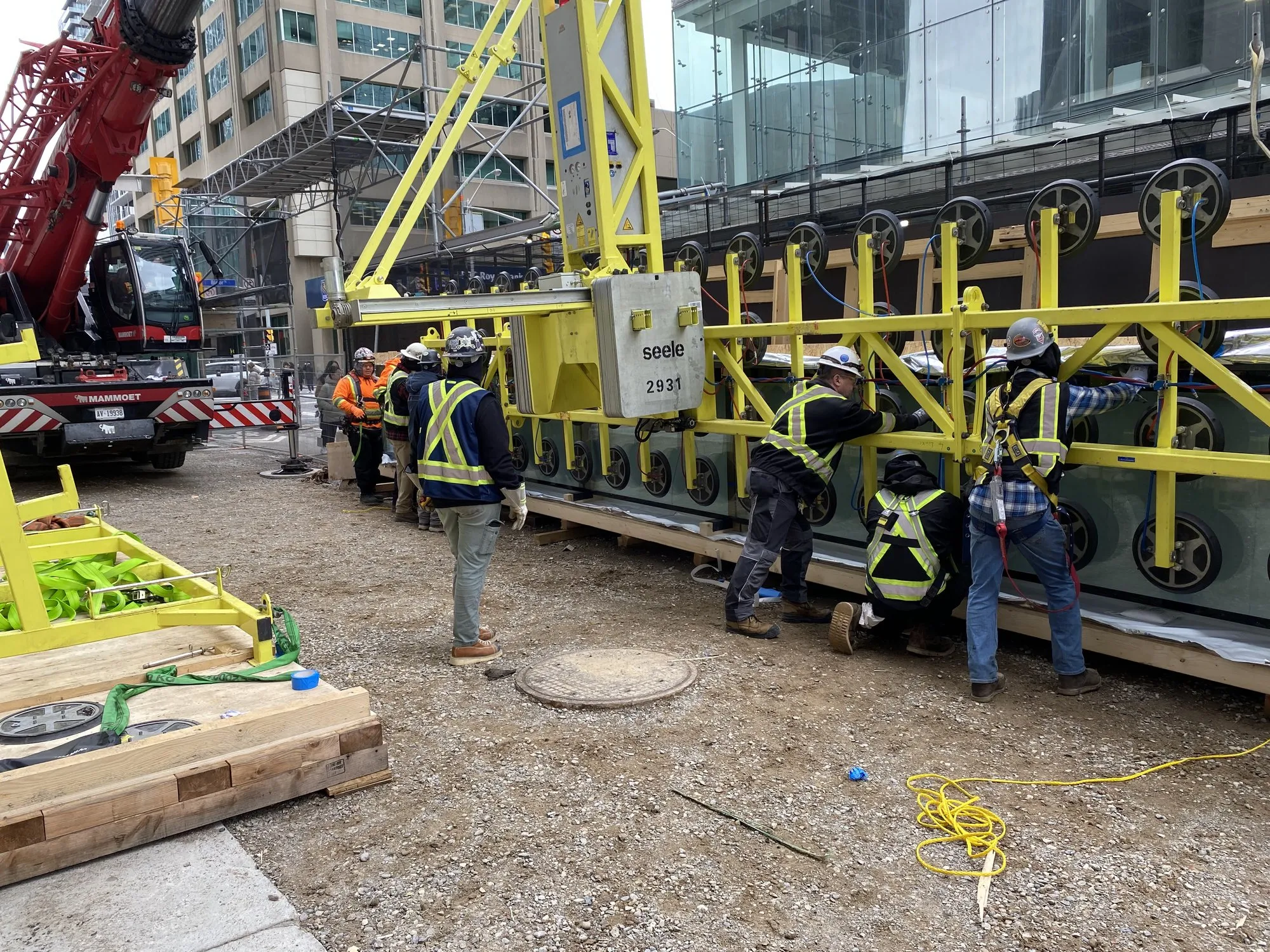A glass panel carried on a vacuum-lifter frame beneath a mobile crane, handled by the installation crew